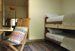 A bunk bed with a patterned pillow, a wooden chair, a small table, and a patterned rug in a room with plain walls.