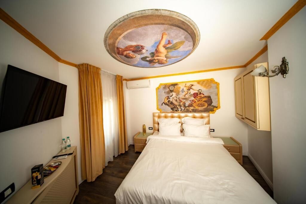 Una habitación de hotel con cama, un mural en el techo, televisor, mesita de noche y ventana con cortinas.