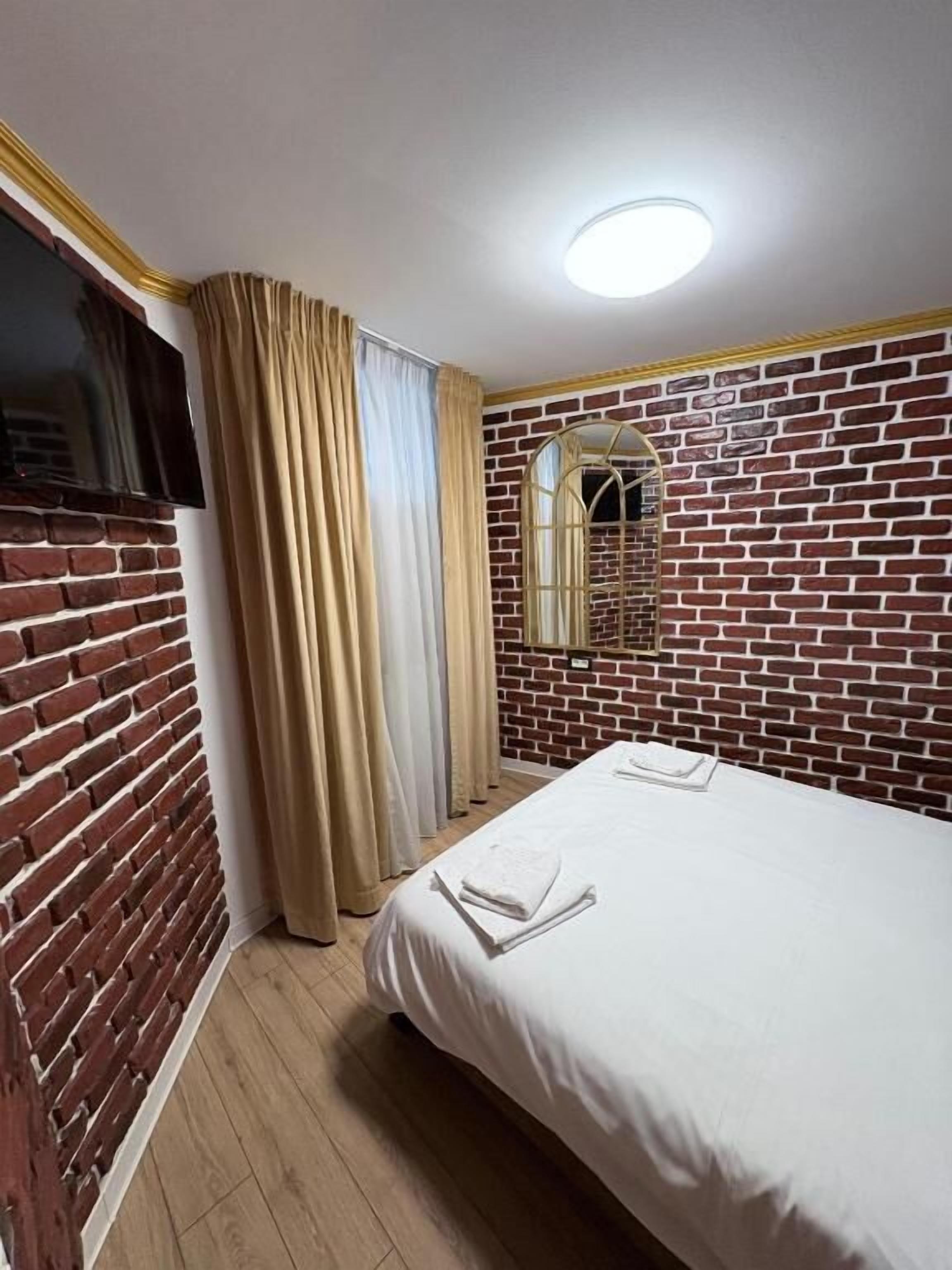 Habitación de hotel con pared de ladrillo, cama con ropa de cama blanca, ventana con cortinas y una lámpara en el techo.