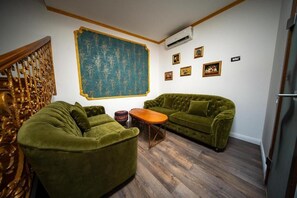 Habitación con sofá verde, una mesa de centro de madera, un tapiz azul grande con motivos y una escalera con barandales ornamentales.
