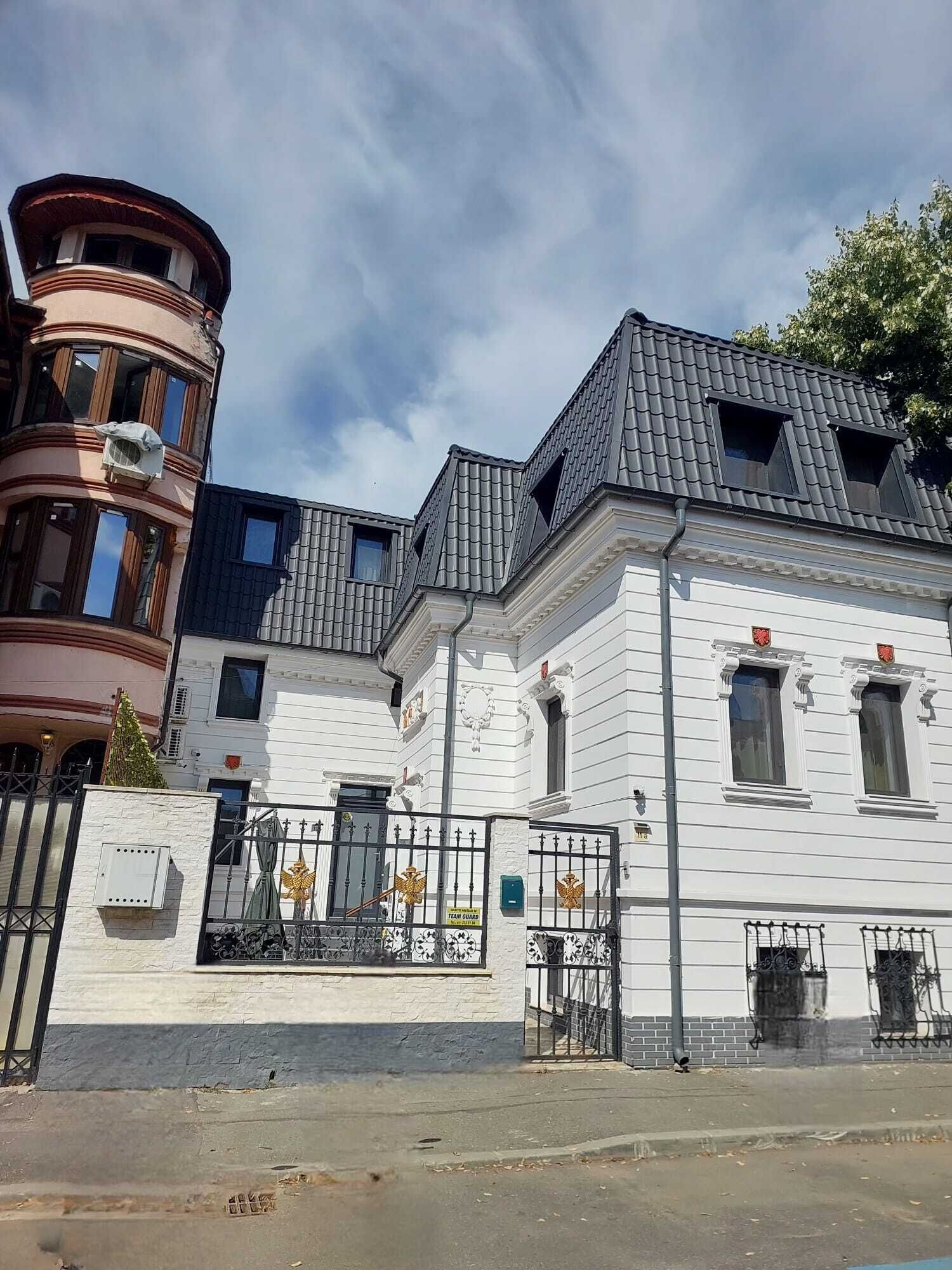 Un edificio con fachada blanca, techo negro y entrada protegida por una reja, con un cartel que indica presencia de seguridad.
