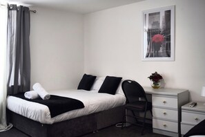 Una habitació d'hotel amb un llit, una taula de treball, una cadira, una tauleta de nit, un vas amb flors i un quadre al mur.