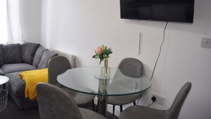 Een moderne woonkamer met een glazen tafel, een grijze bank en een flatscreen televisie aan de muur.