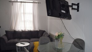Een woonkamer met een bank, een glazen tafel, een afstandsbediening, een bloevas en een wandgemonteerde televisie.