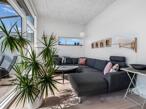 Ein modernes Wohnzimmer mit einem grauen Ecksofa, einem runden Couchtisch und einem Esstisch mit einer Schüssel. Es gibt Topfpflanzen, Kunstwerke an den Wänden und eine Deckenleuchte.
