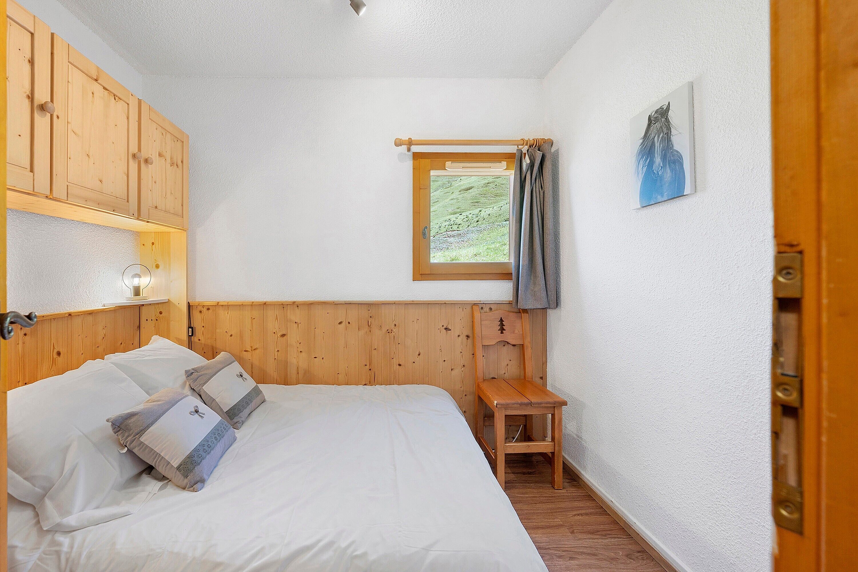 A bedroom with a wooden headboard, a bed with white bedding, a small wooden chair, a window with a view of greenery, and a painting on the wall.