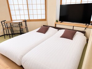 A hotel room with two beds, a TV, a small table with chairs, and a window with a grid pattern. - Newly Opened Right Next to JR Kamata Station: YU'S HOUSE 2 (大田区)