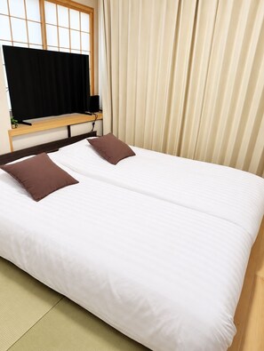 Ein Doppelbett mit zwei Kissen, ein Holz-Kopfteil, ein Fernseher auf einem Schrank und ein Fenster mit Jalousien.