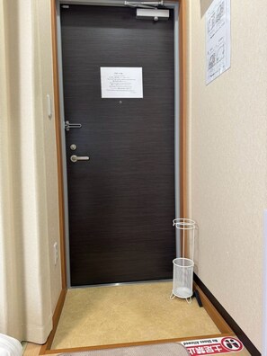 A hotel room entrance with a dark wooden door, a white doormat, and a metal basket on the floor. - Newly Opened Right Next to JR Kamata Station: YU'S HOUSE 2 (大田区)