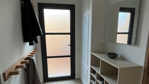 Een gang met een houten kapstok, een deur met geëtste glaspanelen, een wit wandplankje met schoenen en een spiegel.