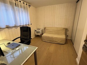 Ein Zimmer mit einem Glastisch, einem schwarzen Bürostuhl, einem beigen Sofa, einem kleinen weißen Nachttisch mit einem Drucker und Holzwandverkleidungen.