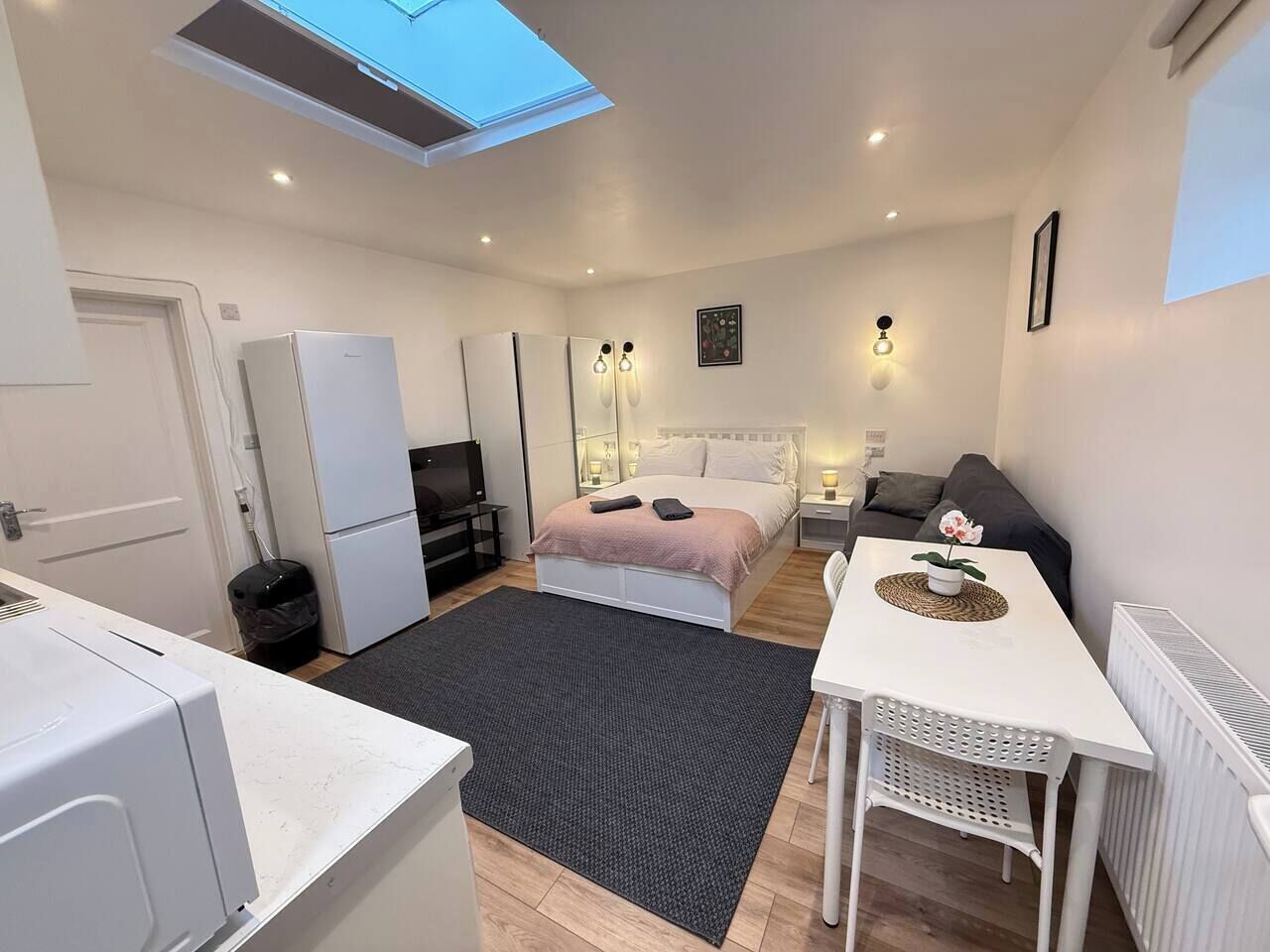 Habitación compacta con cama, pequeña mesa, sofá, kitchenette y tragaluz.