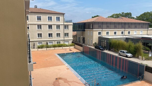 Une piscine avec un plongeoir, un espace abrité et un bâtiment avec de nombreuses fenêtres en arrière-plan.