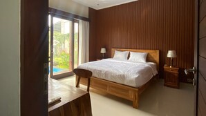 A bedroom with a large bed, a wooden nightstand, a desk, and a view of a swimming pool through a sliding door. - Cozzy Ketewel Beach Villa (gianyar)