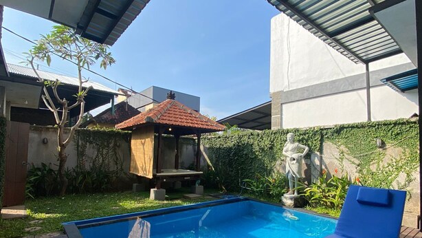 A swimming pool with a wooden deck, a blue lounge chair, and a covered area with a metal roof. - Cozzy Ketewel Beach Villa (gianyar)