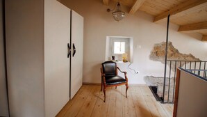 Een kamer met een houten vloer, een zwarte leren fauteuil, een witte kledingkast, een kroonluchter, een raam met uitzicht en een stenen muur.