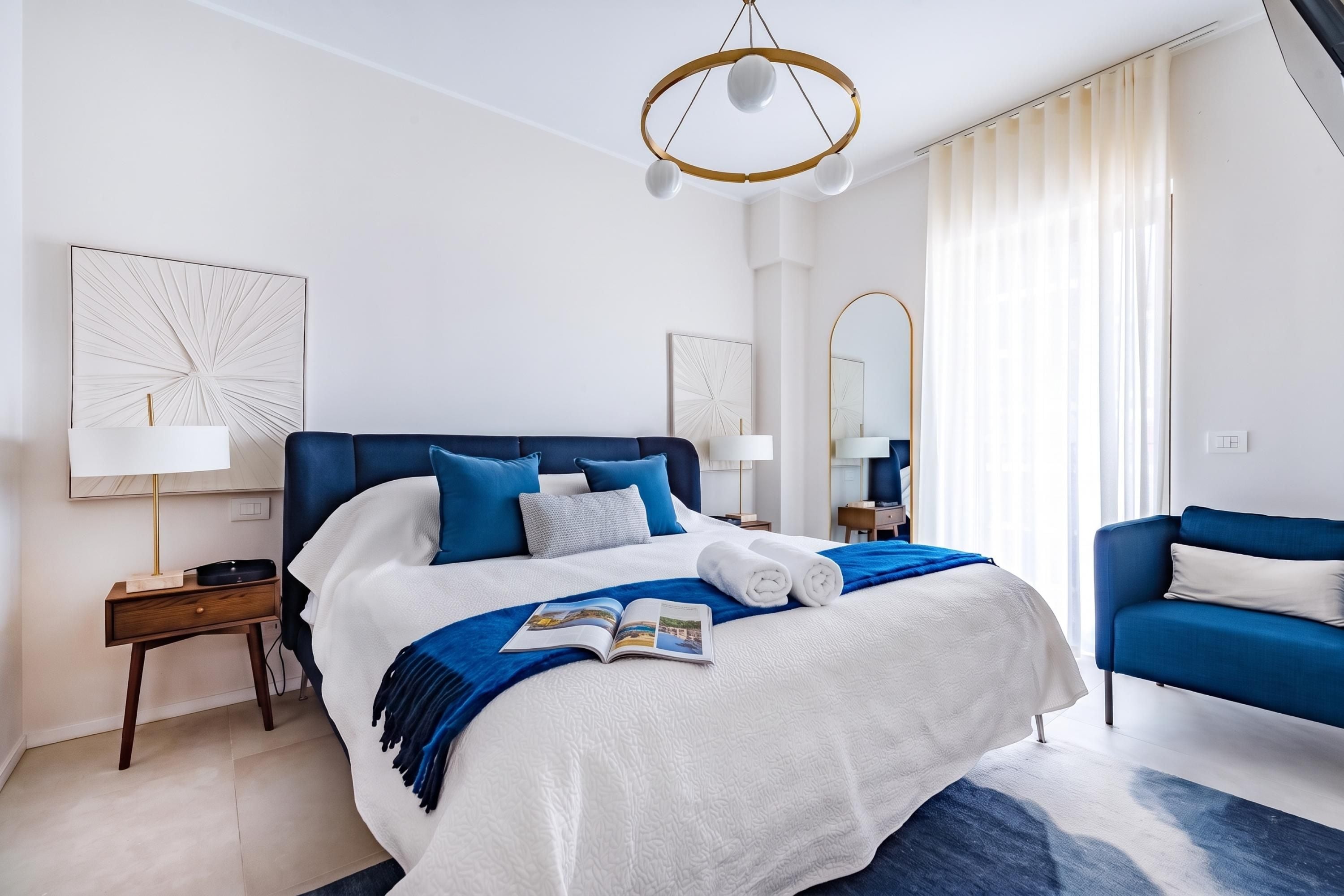 A modern hotel room with a large bed, two bedside tables, a desk, a chair, a large mirror, and a chandelier.