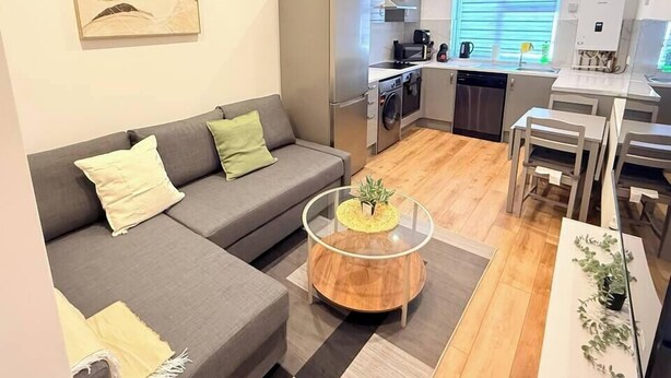 Een modern appartement met een grijze bank, een ronde salontafel, een keuken met een vaatwasser en een eettafel met stoelen.
