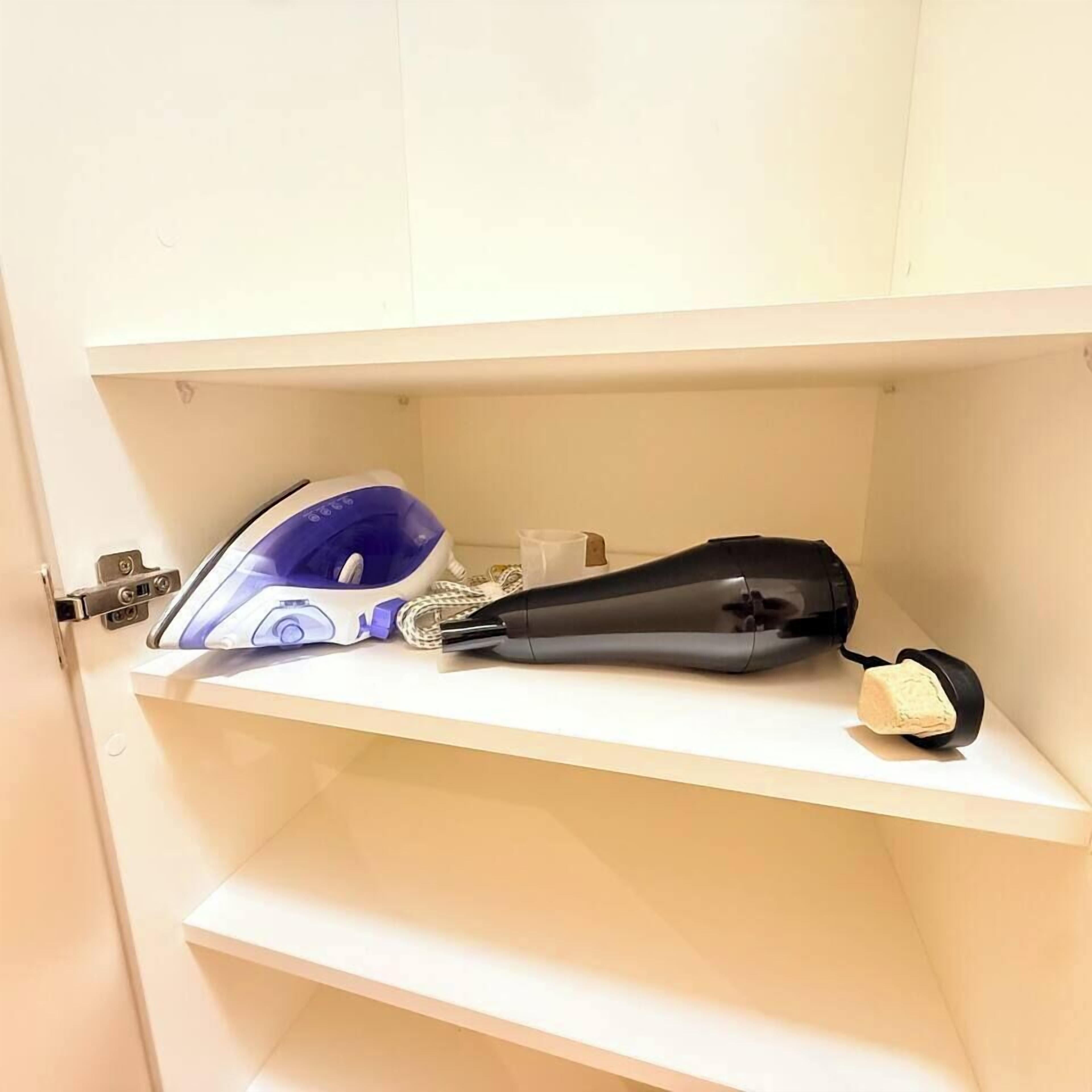 A white shelf with a hairdryer and a blue appliance, possibly a hair straightener, on a white wall.