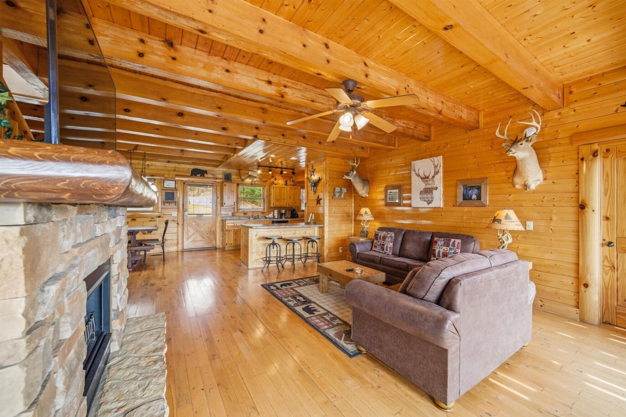 A log cabin interior with a stone fireplace, wooden ceiling beams, a ceiling fan, a kitchen area, and a living room with a sofa and a rug.