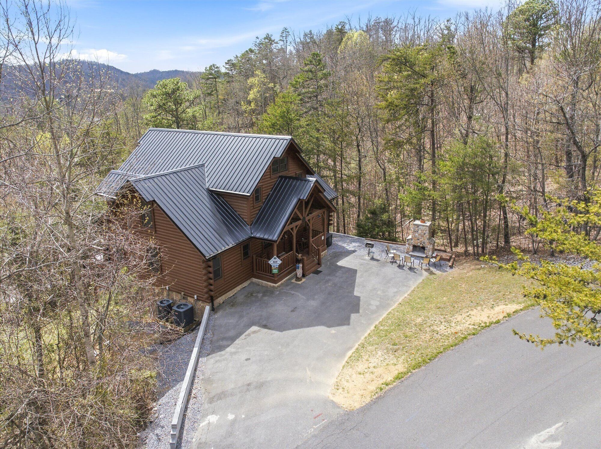 A log cabin with a metal roof, a stone fireplace, and a paved driveway.