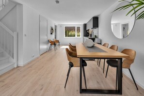A spacious dining area with a long table and chairs, a kitchen in the background, and a staircase on the left.