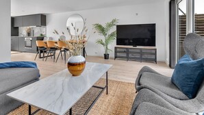 Una sala de estar moderna con una mesa de centro de mármol, sofás grises, un televisor de pantalla plana y un comedor con sillas y una planta.