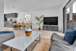 A modern living room with a marble coffee table, grey sofas, a flat-screen TV, and a dining area with chairs and a plant.