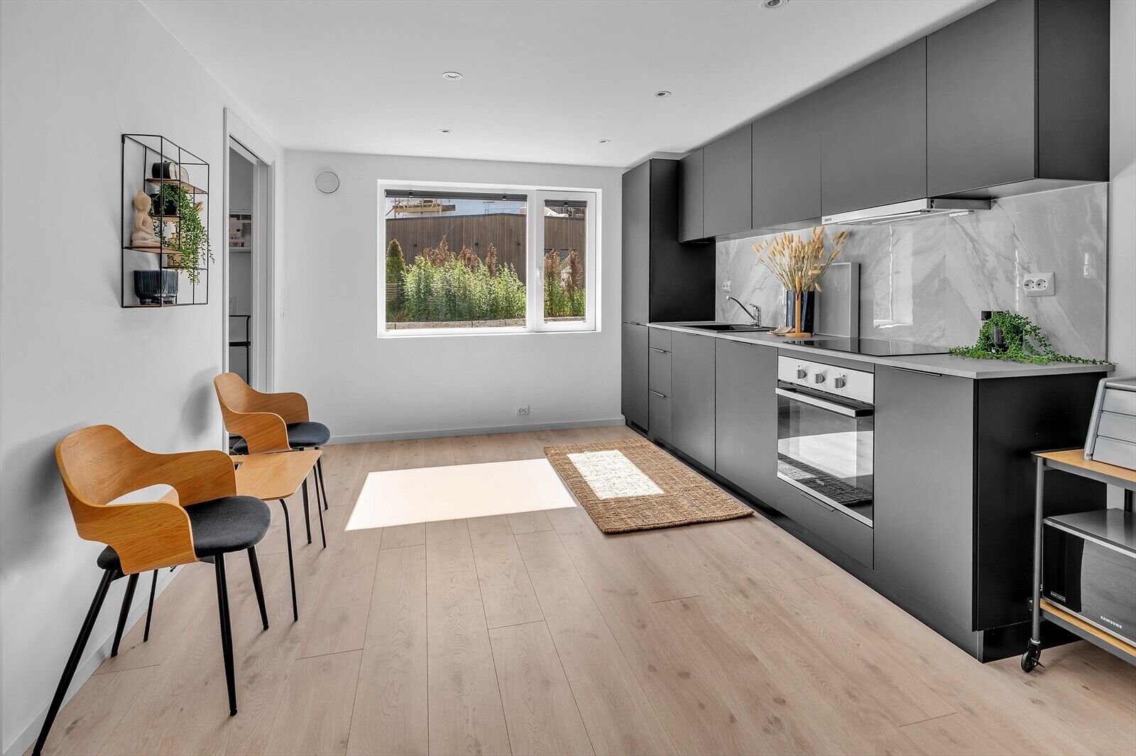 Una cocina moderna con gabinetes grises, horno empotrado y ventana con vistas a un jardín.