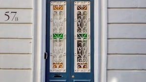 Eine blaue Tür mit einem dekorativen Fensterglas, ein weißer Türrahmen und die Nummer '57B' an der Wand.