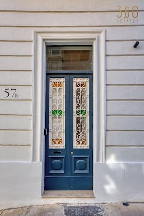 Eine blaue Tür mit einem dekorativen Fensterglas, ein weißer Türrahmen und die Nummer '57B' an der Wand.