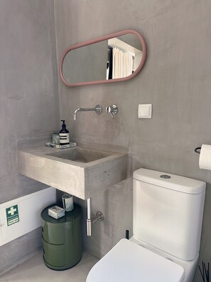 Bany amb un rentamans de formigó, un vàter, un mirall i un kit de primers auxilis.