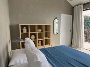 Un dormitori amb llit, una làmpada de nit, un mirall, una estanteria amb objectes decoratius i una vista per una porta.