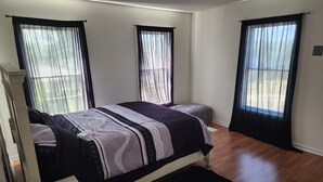 A bedroom with a bed, a bedside table, a window with curtains, and a wooden floor. - The Hideout, a spacious 5BR, 3BT home that's perfect for families friends. (Lanham)