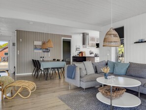 Ein modernes Wohnzimmer mit grauem Sofa, rundem Couchtisch, Korbstuhl, Esstisch mit Stühlen und einer Küche im Hintergrund.