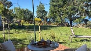 Un espace de restauration en plein air, avec des chaises en osier et une table ronde, entouré d’un paysage verdoyant et d’arbres.