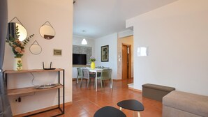 Uma sala de estar com sofá, dois banquinhos pretos, TV, área de refeições com cadeiras e cozinha com fogão.