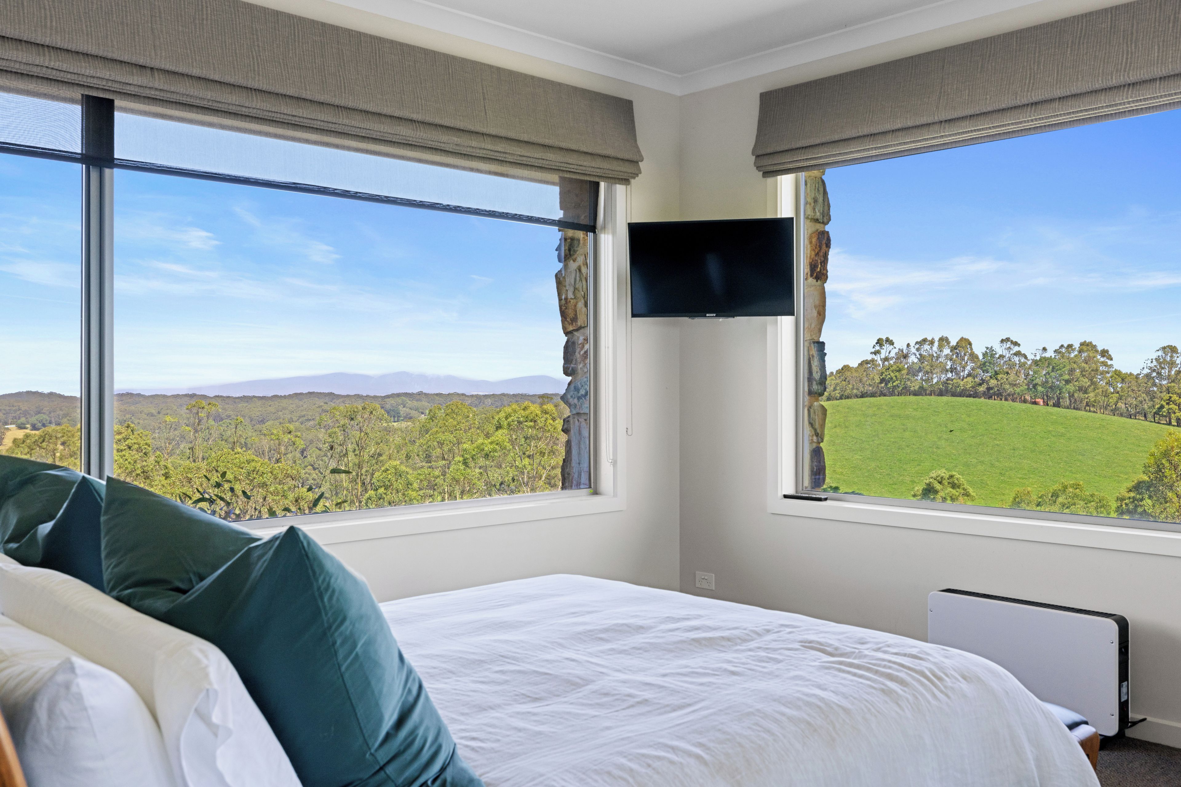 A bedroom with a bed, a flat-screen TV mounted on the wall, and large windows offering a view of a green landscape.
