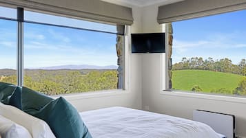 A bedroom with a bed, a flat-screen TV mounted on the wall, and large windows offering a view of a green landscape.