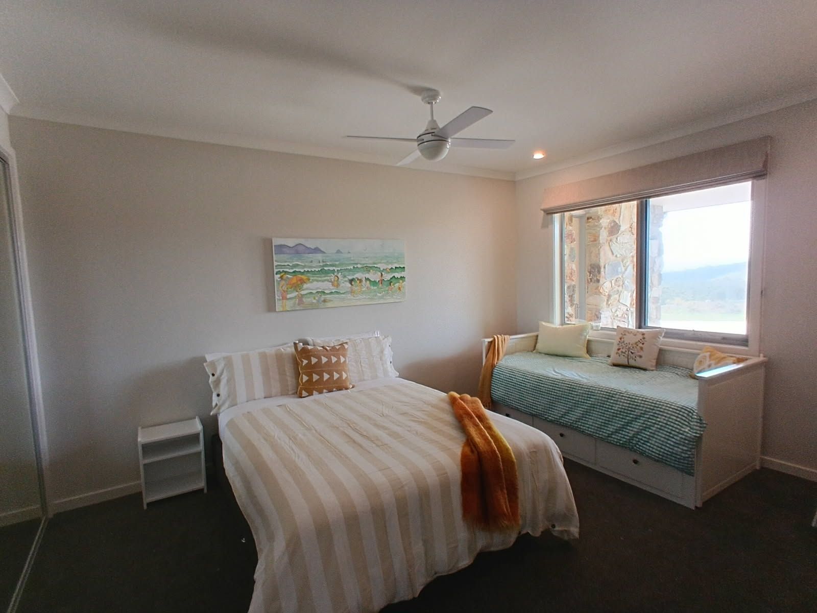 Un dormitorio con cama, un rincón con asiento junto a la ventana, un ventilador de techo y un cuadro en la pared.