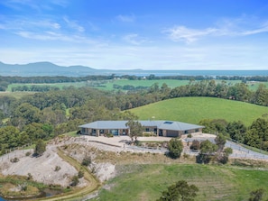 A modern house with a large roof, surrounded by lush green fields and trees, with a clear view of the distant mountains.