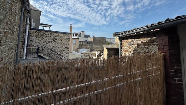 Une cour bordée de bâtiments en pierre, une clôture en bambou et un ciel dégagé.
