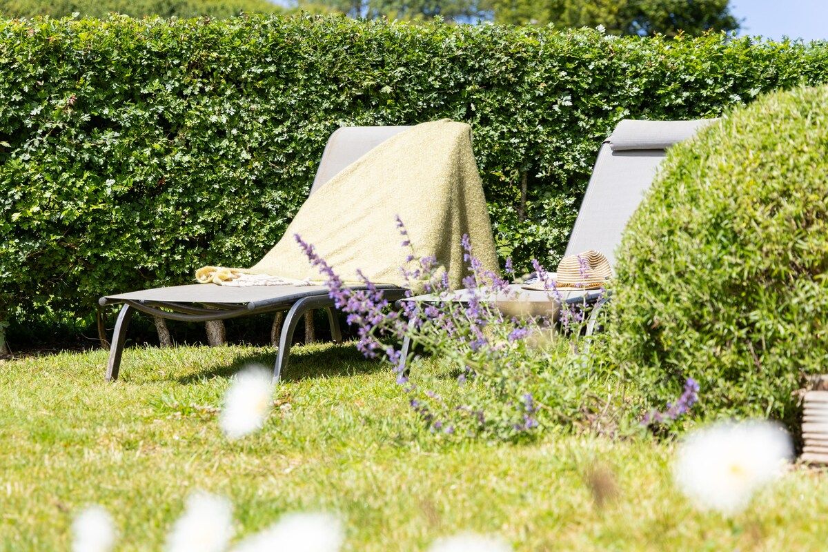 Dos sillones de descanso con una manta, rodeados de follaje y flores púrpuras.