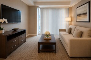 A modern hotel room with a flat-screen TV, a wooden console table with a vase of flowers, a beige sofa, and a large window with blinds. - Mayfairs Hotel (Toronto)