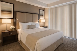 A hotel room with a large bed, two bedside tables, a nightstand, a window with blinds, and two framed pictures on the wall. - Mayfairs Hotel (Toronto)