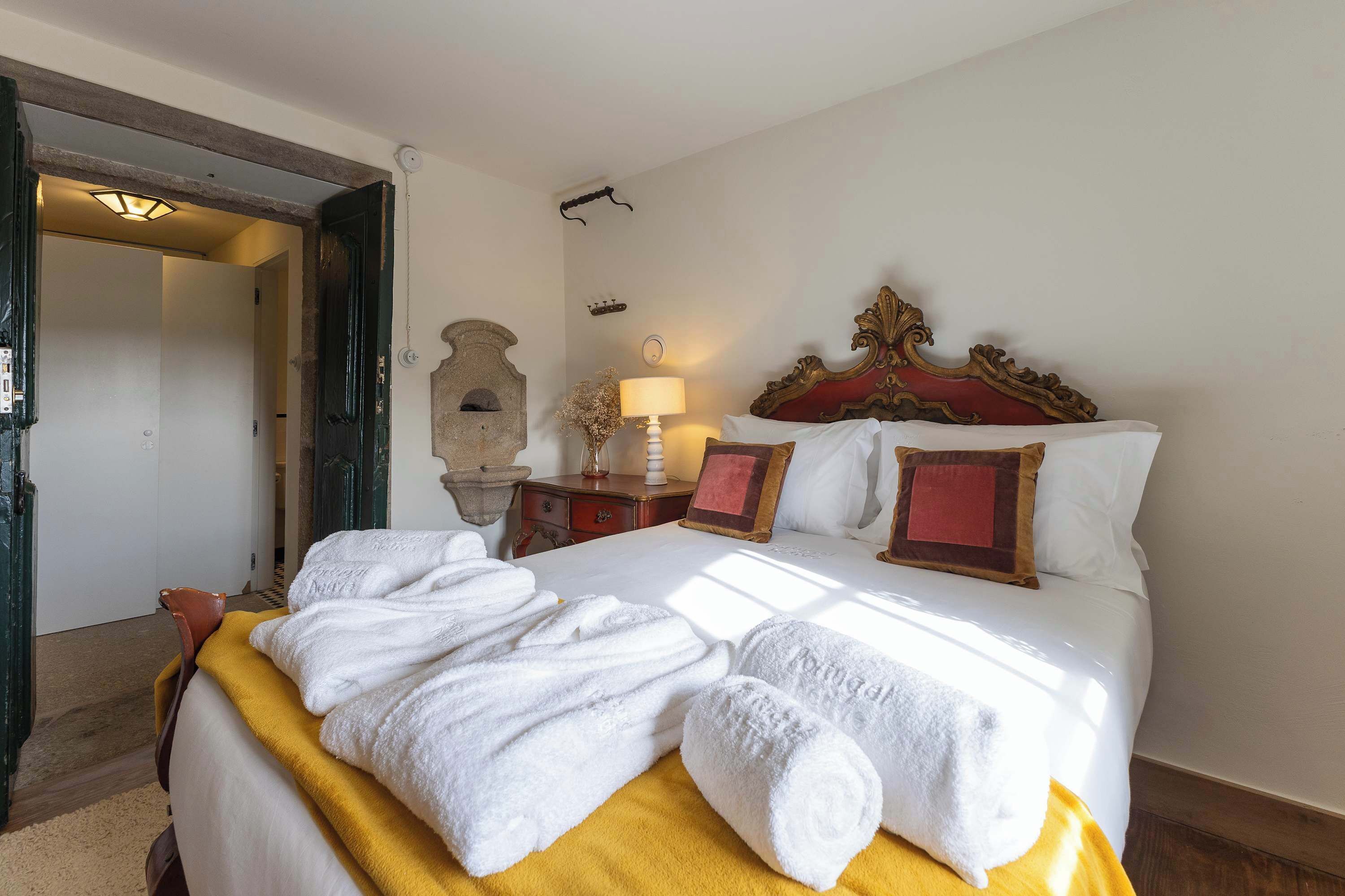 A hotel room with a bed, a nightstand, a lamp, and a door leading to another room.