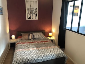 Ein ordentlich bezogenes Bett mit gemusterten Bettwäsche, zwei Nachttischlampen, eine Wanduhr und ein gerahmtes Bild des Empire State Buildings.