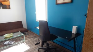 Ein Zimmer mit braunem Sofa, einem Glastisch mit einem Buch, einem schwarzen Bürostuhl, einem Schreibtisch mit Computer und einem gerahmten Bild an der Wand.