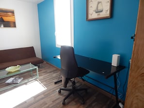 Ein Zimmer mit braunem Sofa, einem Glastisch mit einem Buch, einem schwarzen Bürostuhl, einem Schreibtisch mit Computer und einem gerahmten Bild an der Wand.
