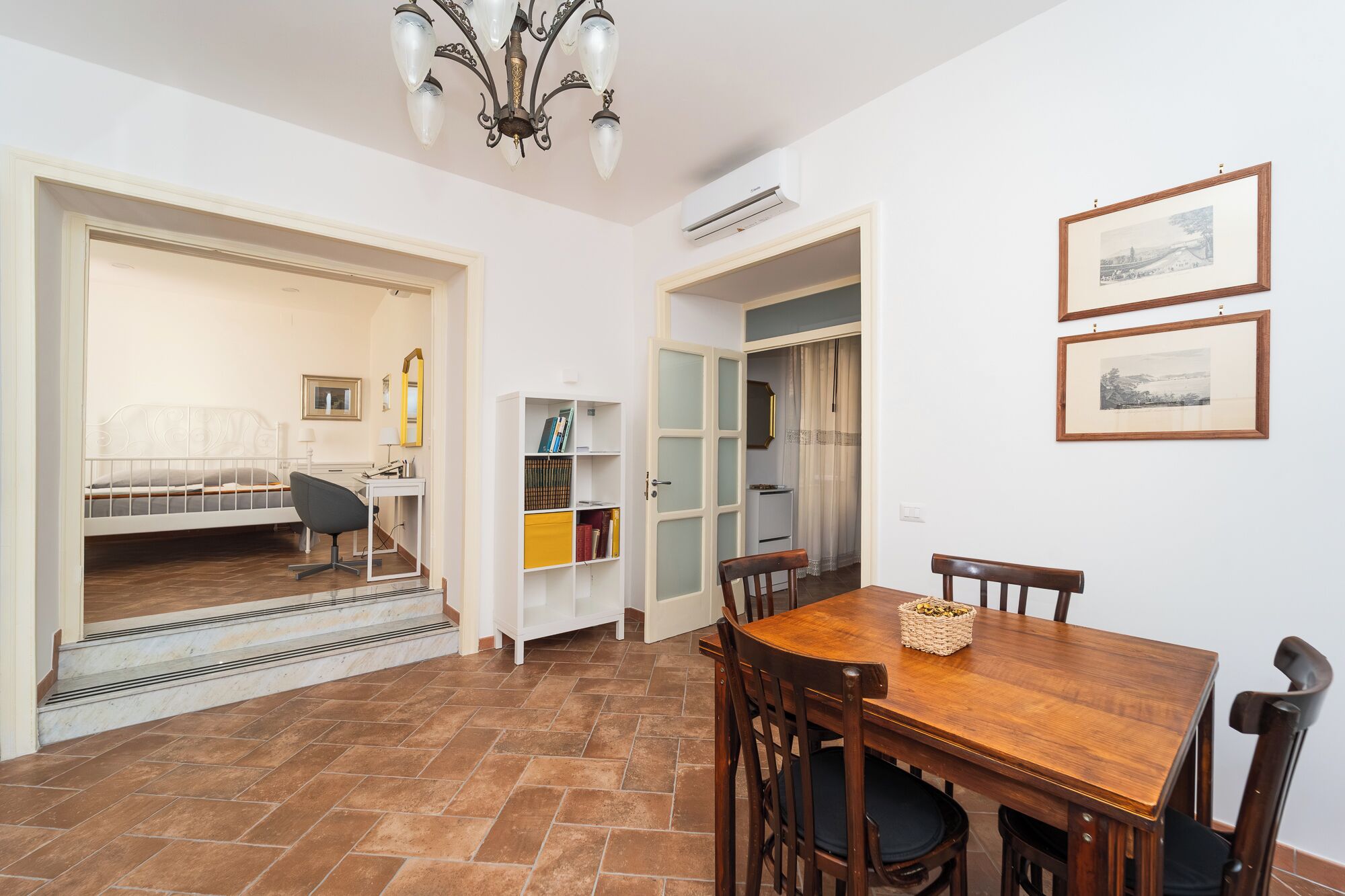 A room with a bed, a desk, a chair, a dining table with chairs, a bookshelf, and two framed pictures on the wall.